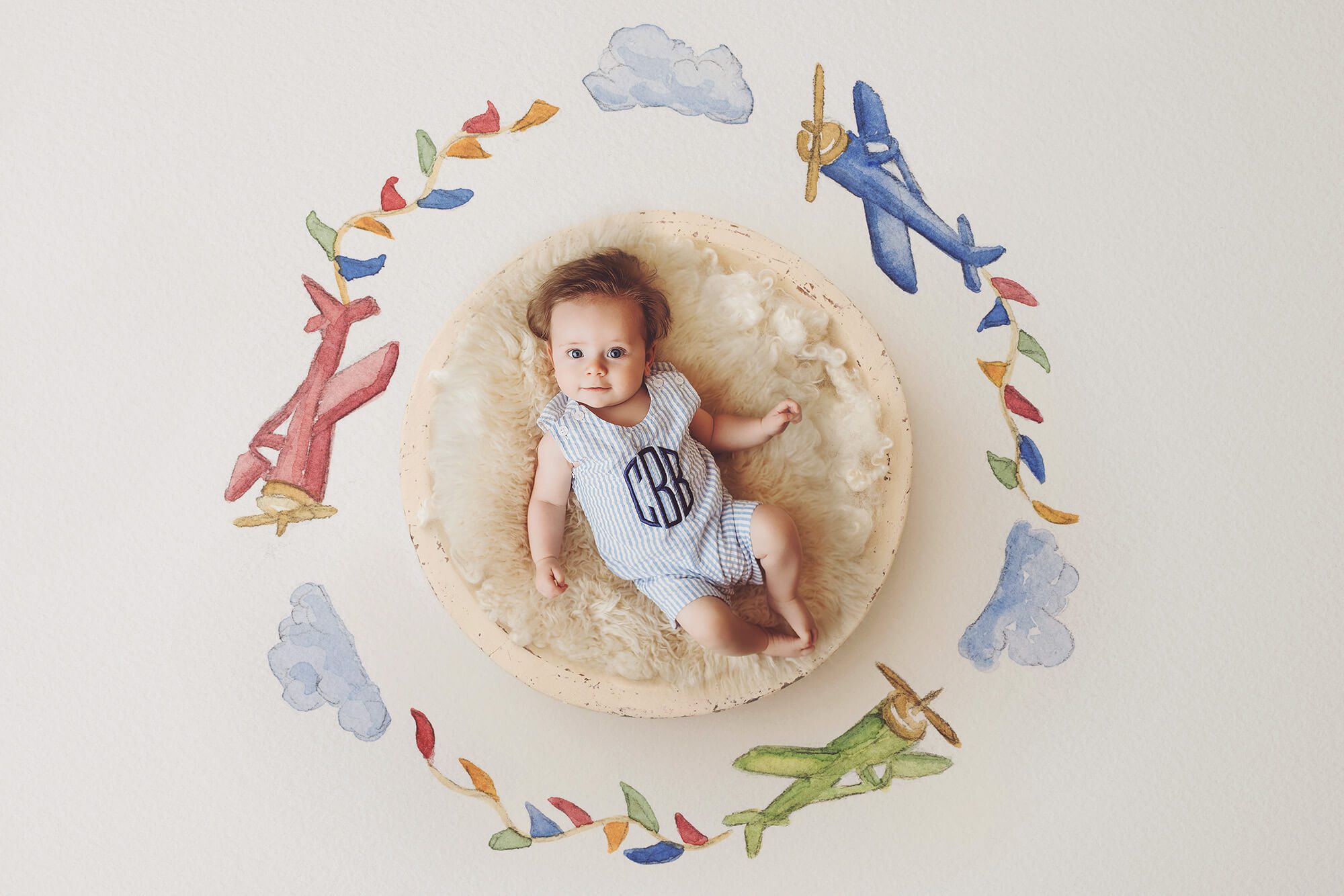 A three-month old baby boy laying in a wooden bowl surrounded by a backdrop of hand-drawn airplanes