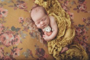 newborn girl holding a bear laying on a yellow floral blanket by clay Kaserne newborn photographer