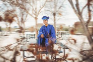 A senior at Catalina Foothills High School sits on the benches of his school, while the world bustles around him wearing his cap and gown