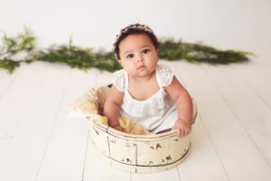 Baby girl in a cream bucket with a white dress and greenery adorned studio during her 6-month milestone session in the Belle Vie Photography studio