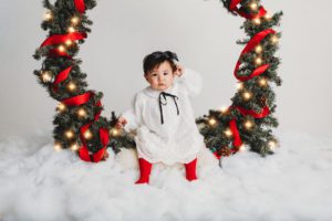 A little girl dressed in her holiday best sits in a giant Christmas wreath during holiday photo sessions with Belle Vie Photography