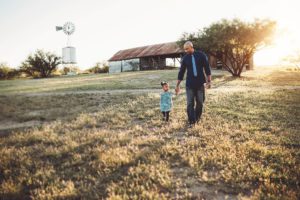 A father and daughter walk in the light of the setting sun with a barn and windmill behind them