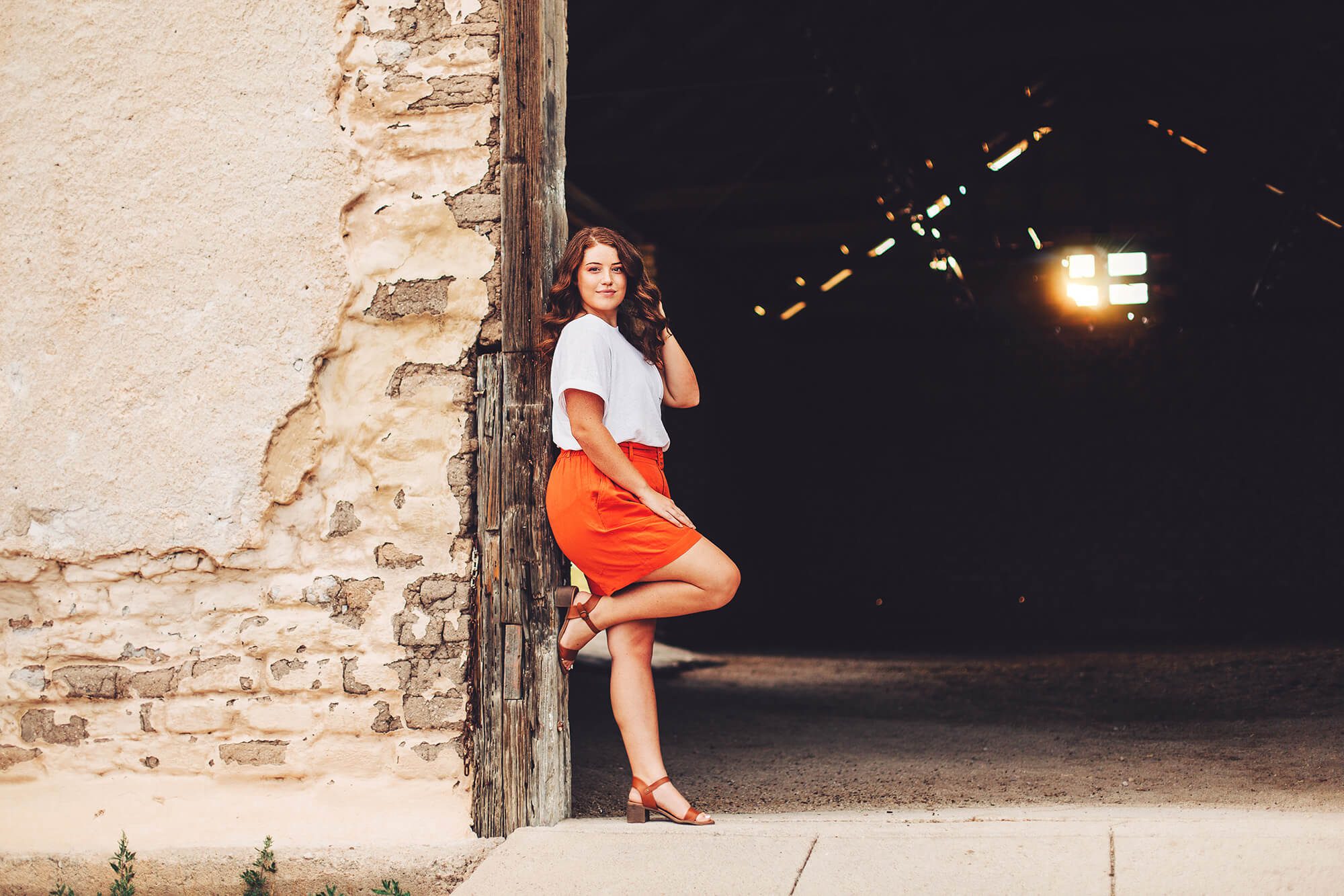 A senior girl leans against a barn entrance playing with her hair as the sun shines through the barn window during her senior session in Sonoita with Belle Vie Photography