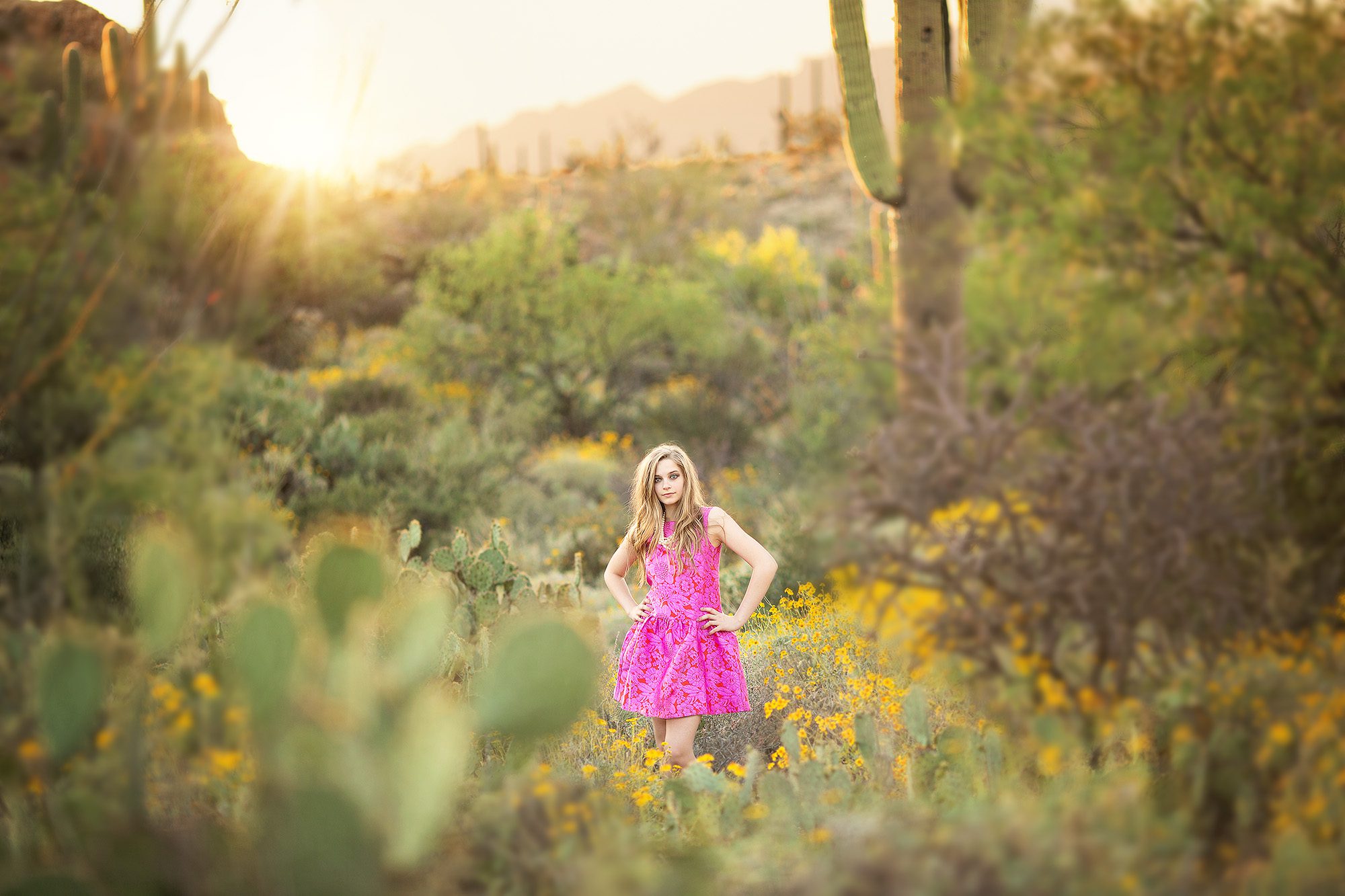 Teenage girl wearing a colorful pink dress and pearls during her photo session at Saguaro National Park East, posed amongst saguaros and desert flowers with the Santa Catalina Mountains as a backdrop.
