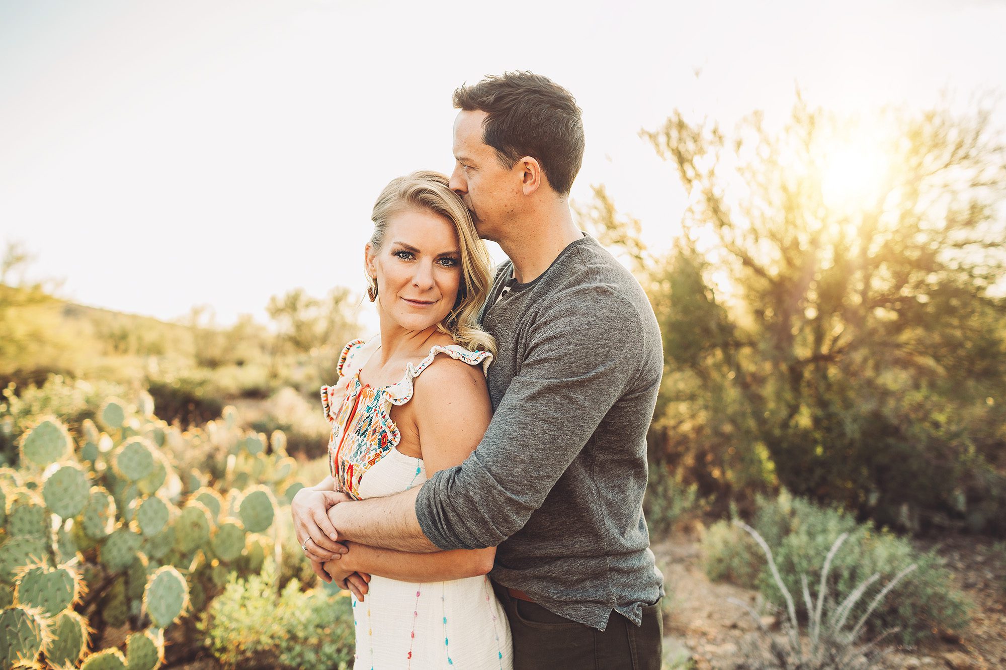 A husband affectionately kisses his wife on the head as they snuggle in the setting sun during their family session at Gates Pass in Tucson