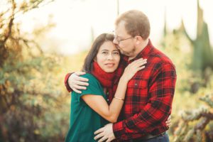 A couple snuggles together for a kiss surrounded by sun and desert foliage during their 2018 holiday photo session with Belle Vie Photography