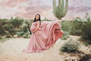 A pregnant mom's dress is artfully thrown into the wind surrounded by green cactus, a giant saguaro and pastel skies during her maternity session in Tucson with Belle Vie Photography