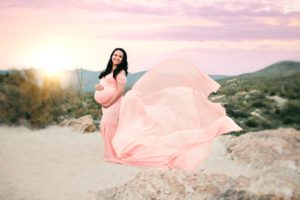 A Tucson mom beautifully poses with her peach maternity dress swirling behind her, the setting sun and catalina mountains at her back, during her sunset maternity session with Belle Vie Photography at Saguaro National Park