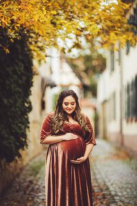 A mom cradling her belly surrounded by fall leaves standing on the cobblestone street in Hochheim