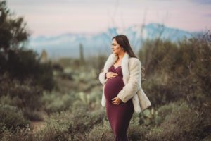 Mom-to-be Anica cradles her baby bump as she gazes at a Tucson sunrise with majestic desert mountains behind her during her maternity session with Belle Vie Photography