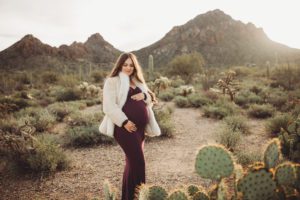 Mom-to-be Anica gazes at her bump amongst prickly pear cactus with rocky Tucson mountains and the rising sun behind her during her maternity session with Belle Vie Photography