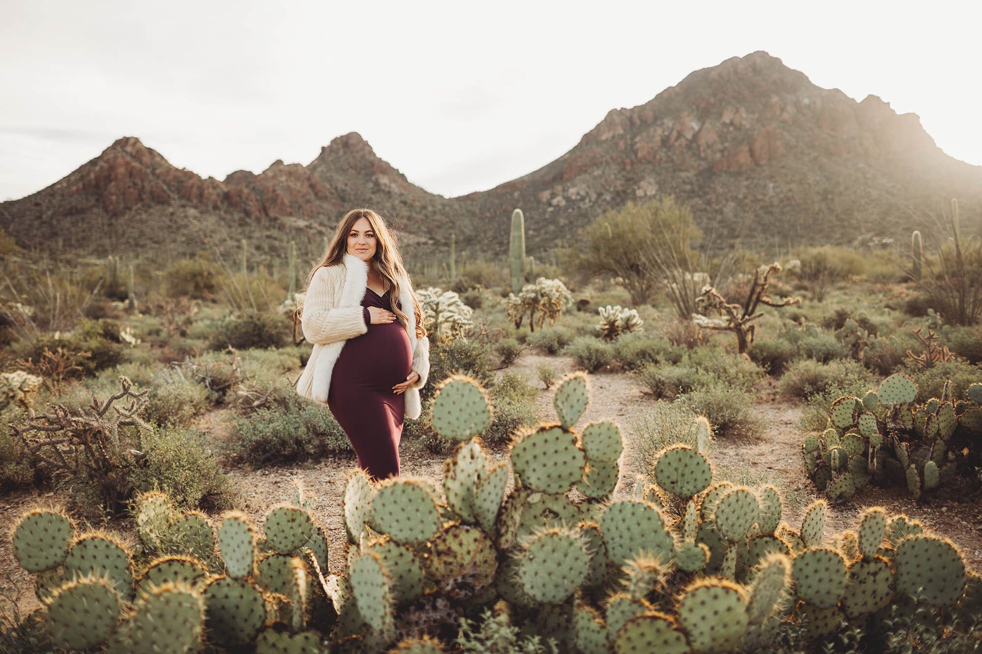 The sun rises over Gates Pass beautifully illuminating Anica and the surrounding cactus during her desert maternity sunrise session with Belle Vie Photography in Tucson