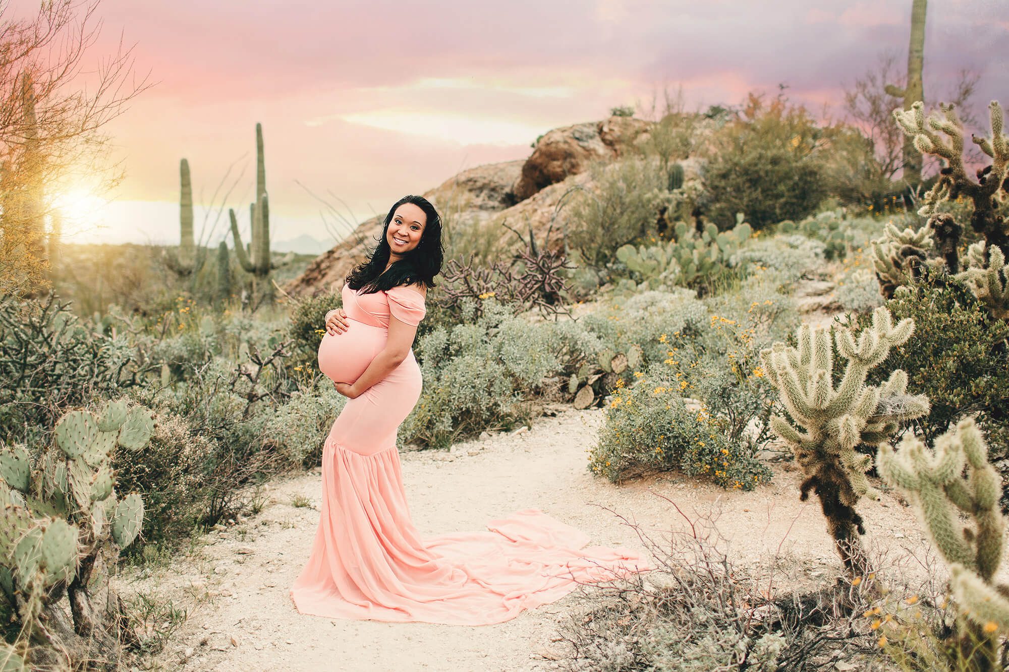 Christina stands amongst the rocks and cacti glowing in her peach gown with a brilliant Tucson sunset behind her