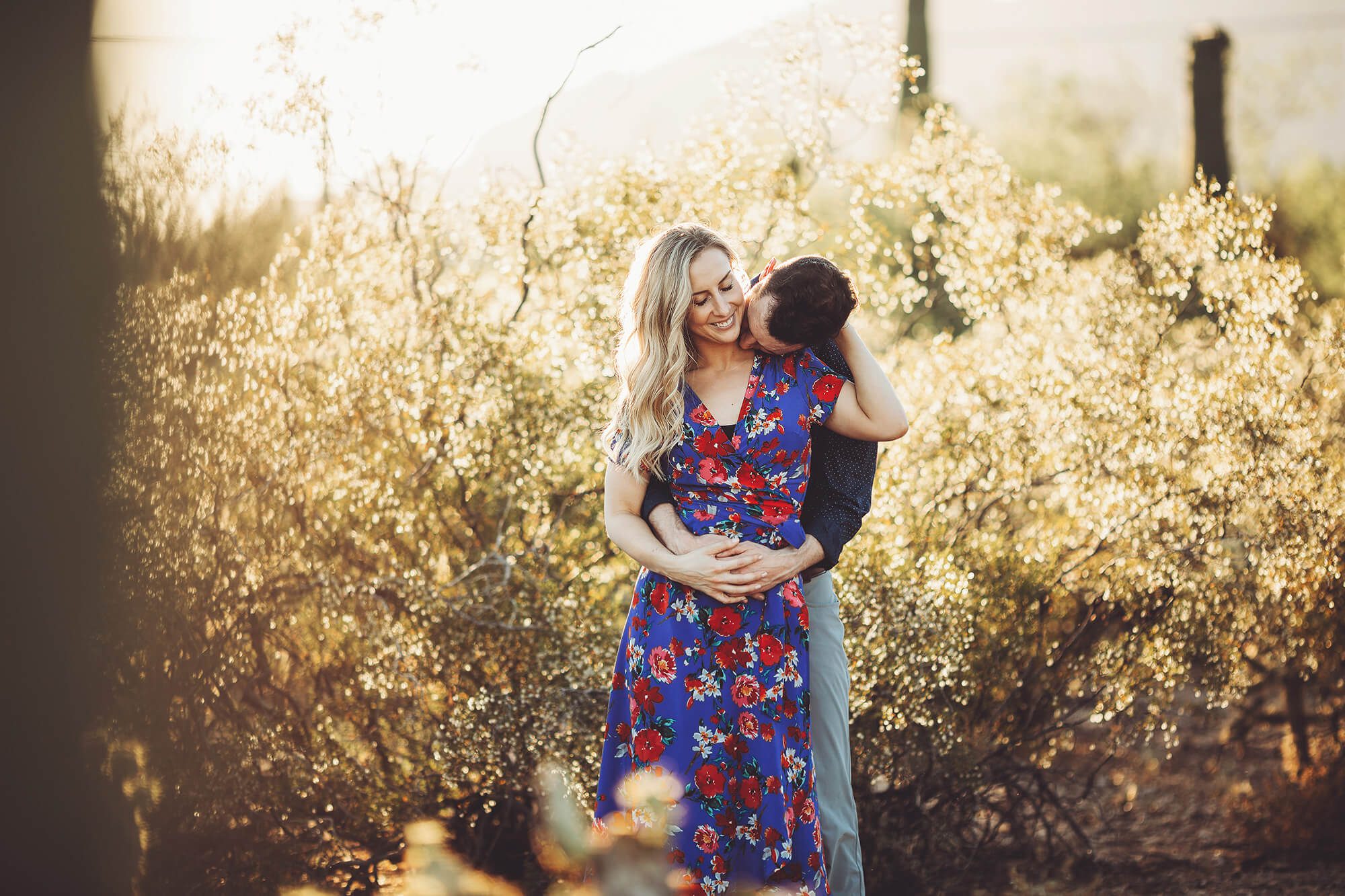 Shaun kisses Ally's neck surrounded by spring desert plants during their sunset engagement session