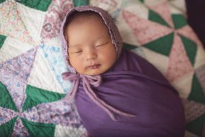 This baby girl looks amazing in a purple wrap and bonnet on a pastel spring quilt