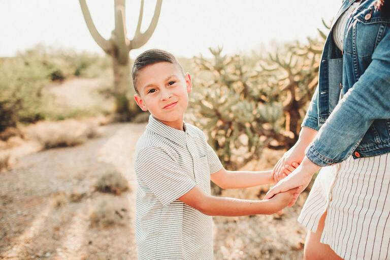 A little boy holding his mom's hands with the desert sun and saguaros at their backs.