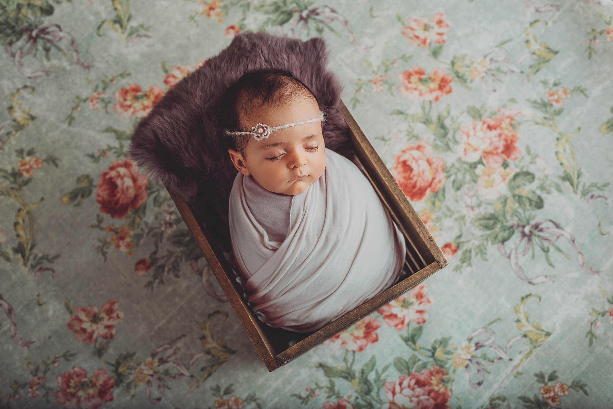 Newborn baby Amelia wrapped in a lavender wrap in a wooden crate with lavender fur on a pink floral backdrop