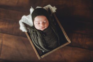 A newborn boy wrapped in sage green in a wooden crate on a wooden floor